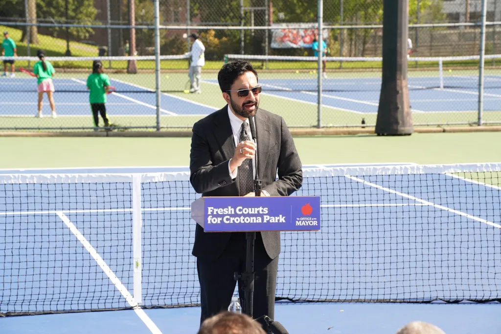Mayor Zohran Mamdani speaking at a podium on a tennis court, with a sign that says 