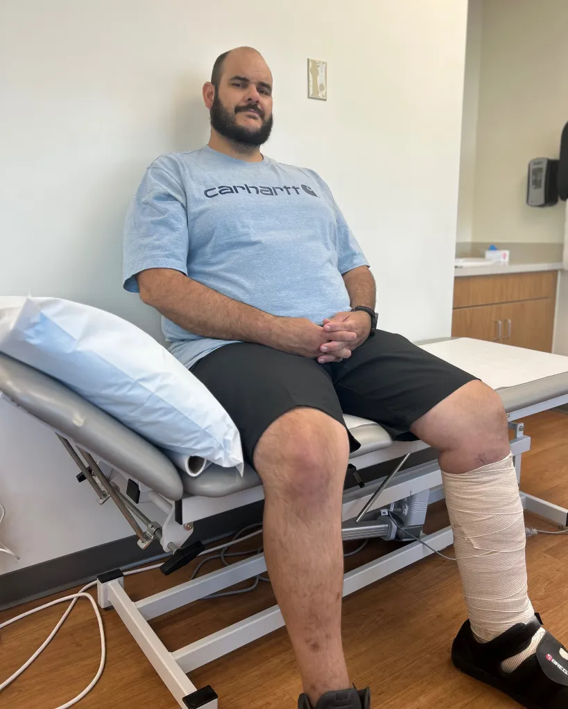 Ben West, who contracted flesh-eating bacteria, sits on an examination table with one leg wrapped in bandages.