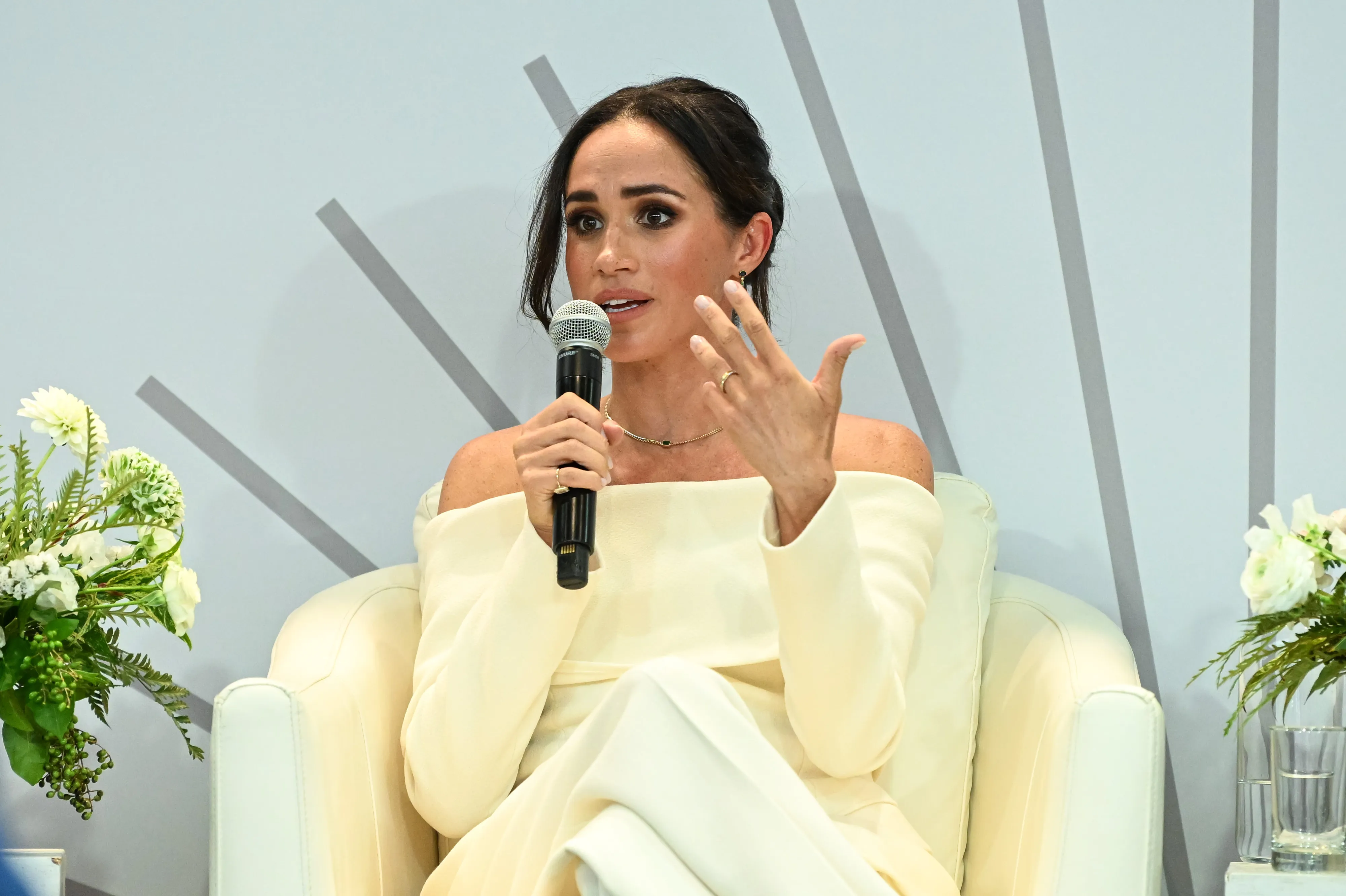 Meghan Markle speaking into a microphone, gesturing with her left hand, while seated in a white chair.
