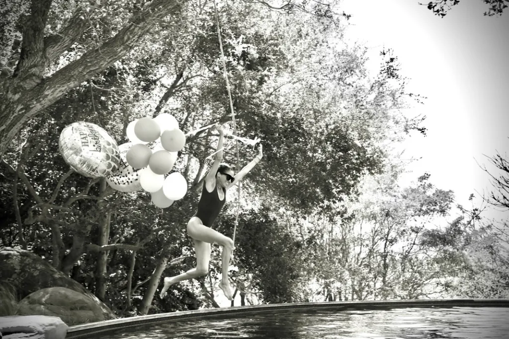 Meghan Markle swinging from a rope into a pool, with balloons nearby.
