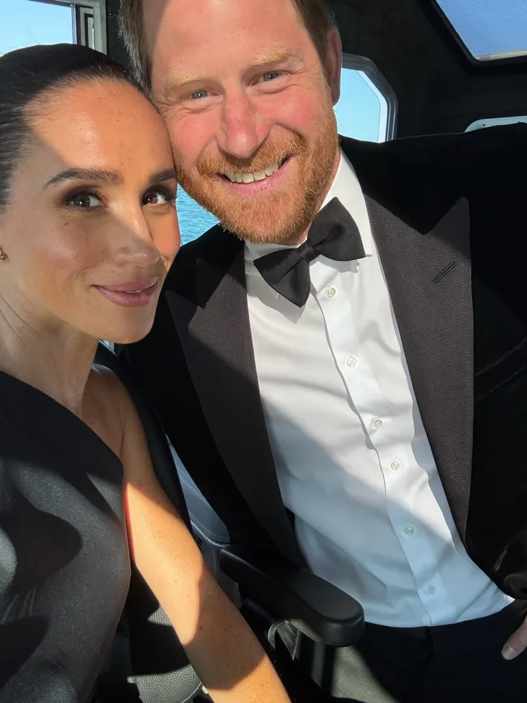 Meghan Markle and Prince Harry pose for a selfie, smiling, with Harry wearing a tuxedo and Meghan wearing a black dress.
