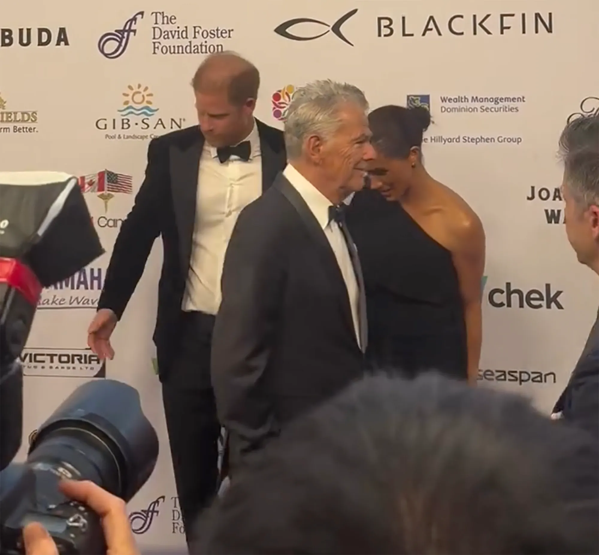 Prince Harry, Meghan Markle, and David Foster on a red carpet with sponsor logos in the background.