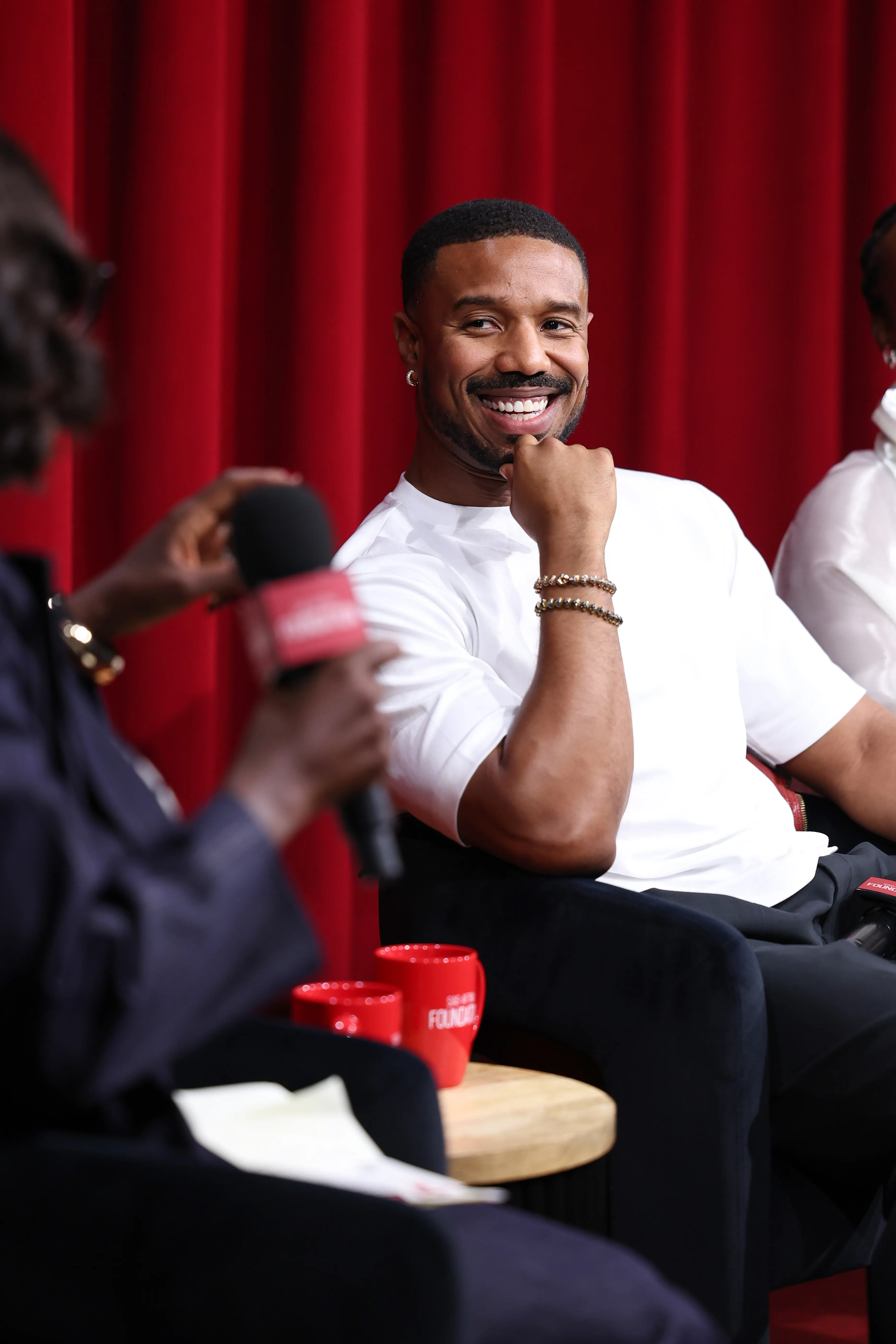 Michael B. Jordan smiling and holding a microphone, with a red curtain in the background.