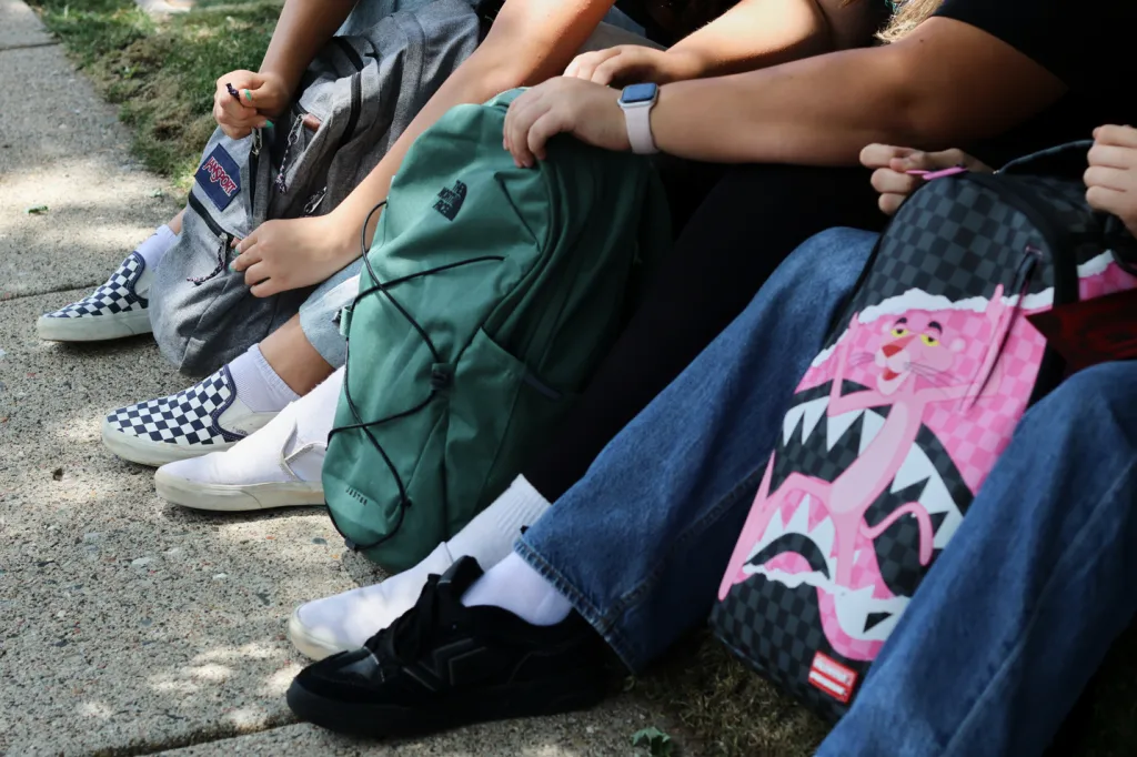 Three students sitting with their backpacks: a gray JanSport, a green North Face, and a black and pink Pink Panther bag.
