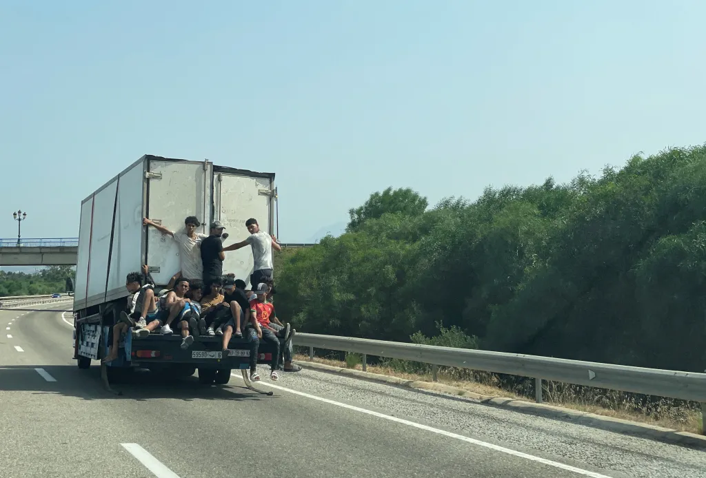Migrants riding on the back of a truck, clinging to the sides, on a highway in Fnideq, Morocco.