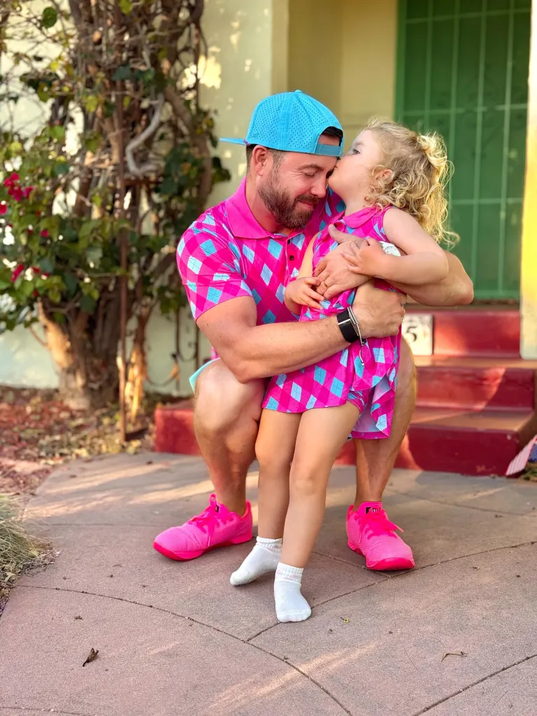 A dad in a bright pink and blue argyle shirt and hat with a little girl in a matching dress, kneeling and embracing, as she kisses his cheek.
