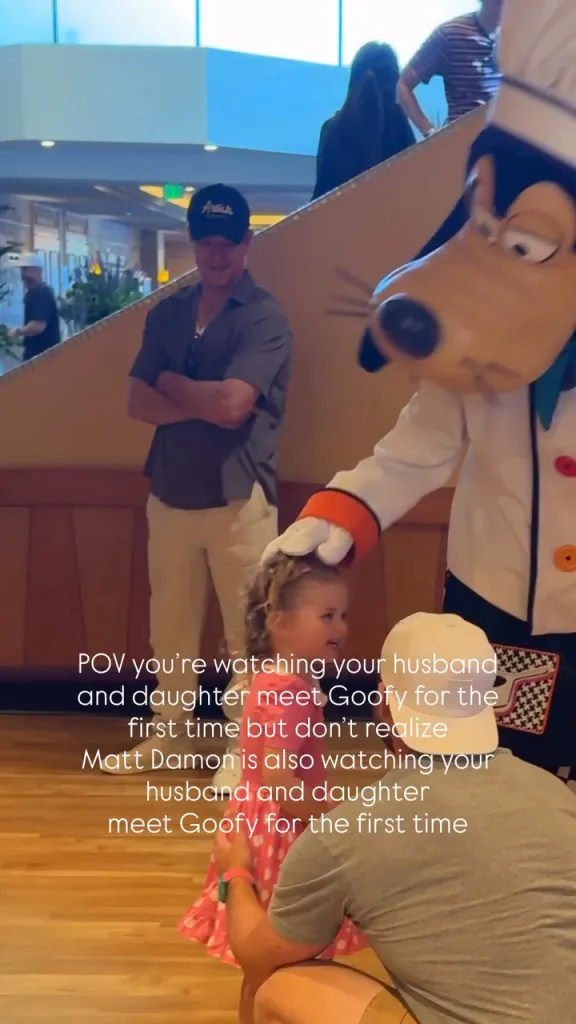 A man in a hat, identified by overlay text as Matt Damon, smiles in the background as Goofy interacts with a child and an adult in the foreground.