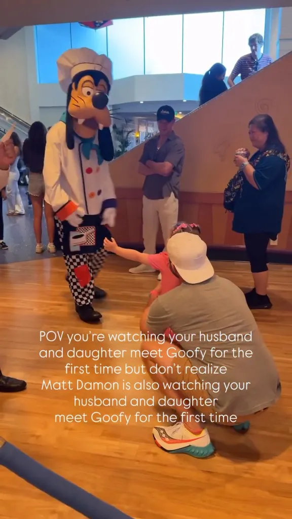 Goofy the Disney character, dressed as a chef, interacting with a young girl and her father at Disneyland, while Matt Damon stands in the background.