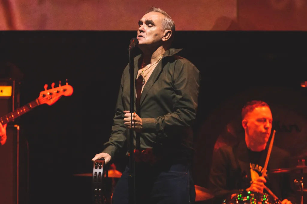 Morrissey sings into a microphone, holding a tambourine, while Matt Walker plays drums in the background.