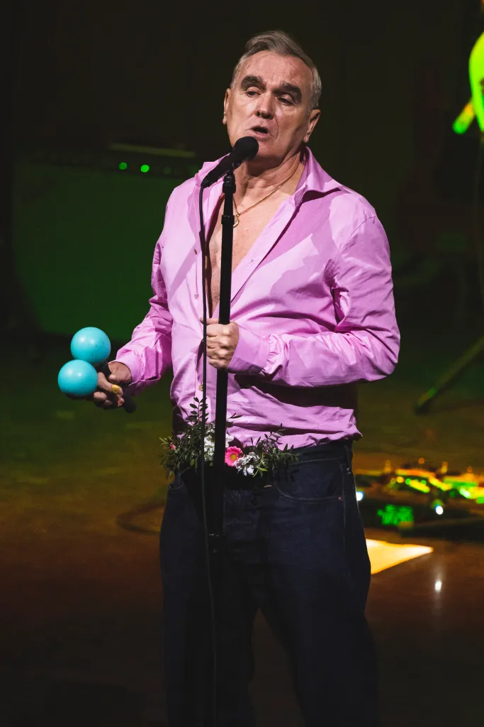Morrissey performing on stage, wearing an unbuttoned pink shirt and blue jeans, holding maracas.