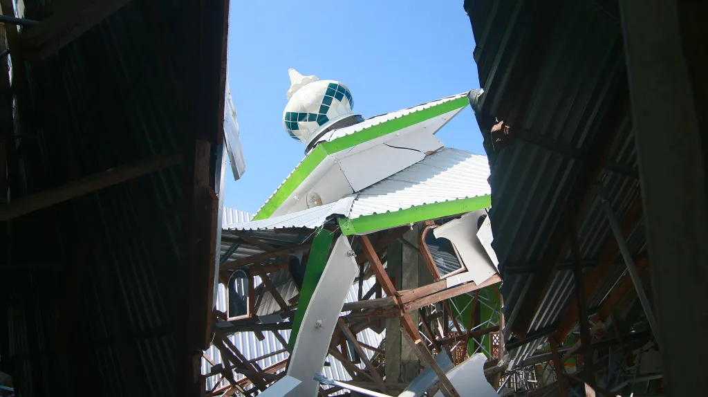 A mosque with a white and green dome roof is destroyed by an earthquake, with its interior wooden beams and metal sheets exposed.