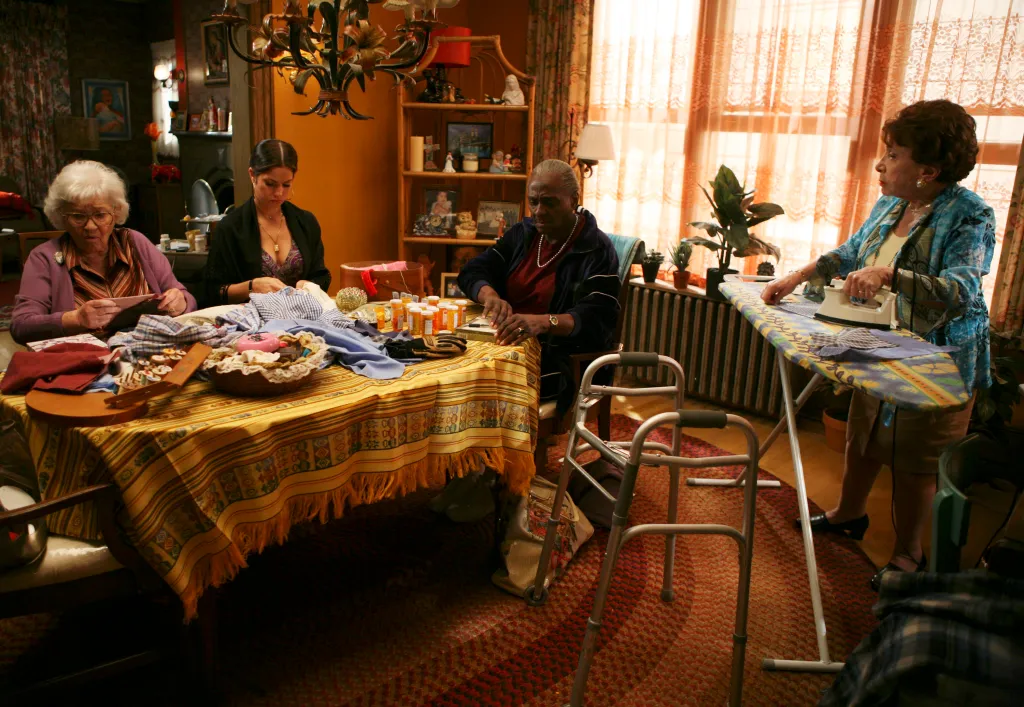 Four women in a living room, one ironing, one at a table with a walker nearby, and two others sitting at the table doing needlework.