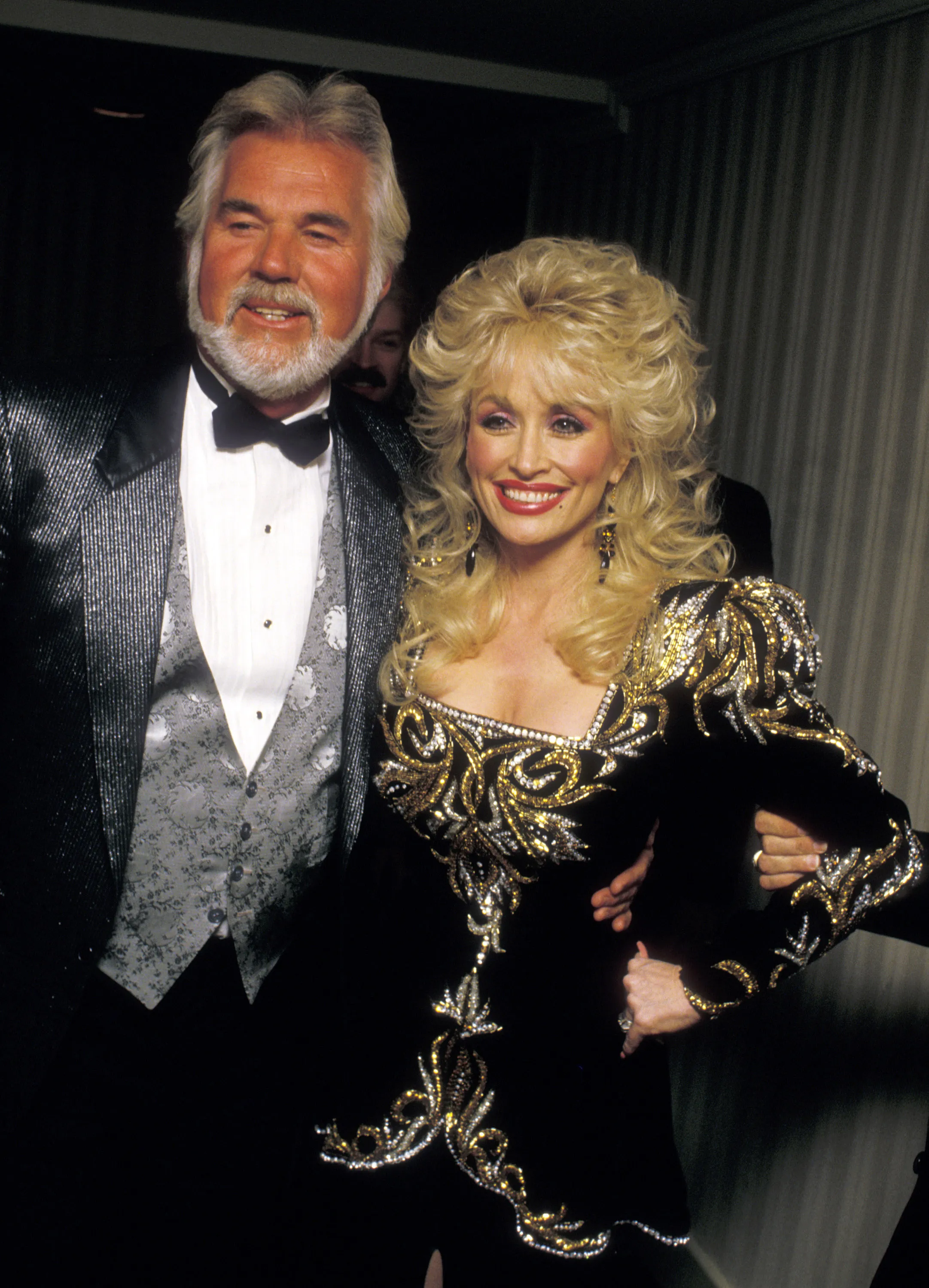 Musicians Kenny Rogers and Dolly Parton smiling and posing for a picture at an event.
