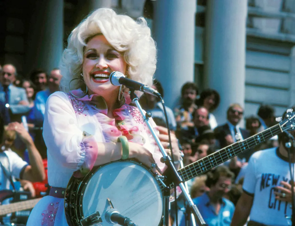 Dolly Parton smiling and singing into a microphone while holding a banjo.