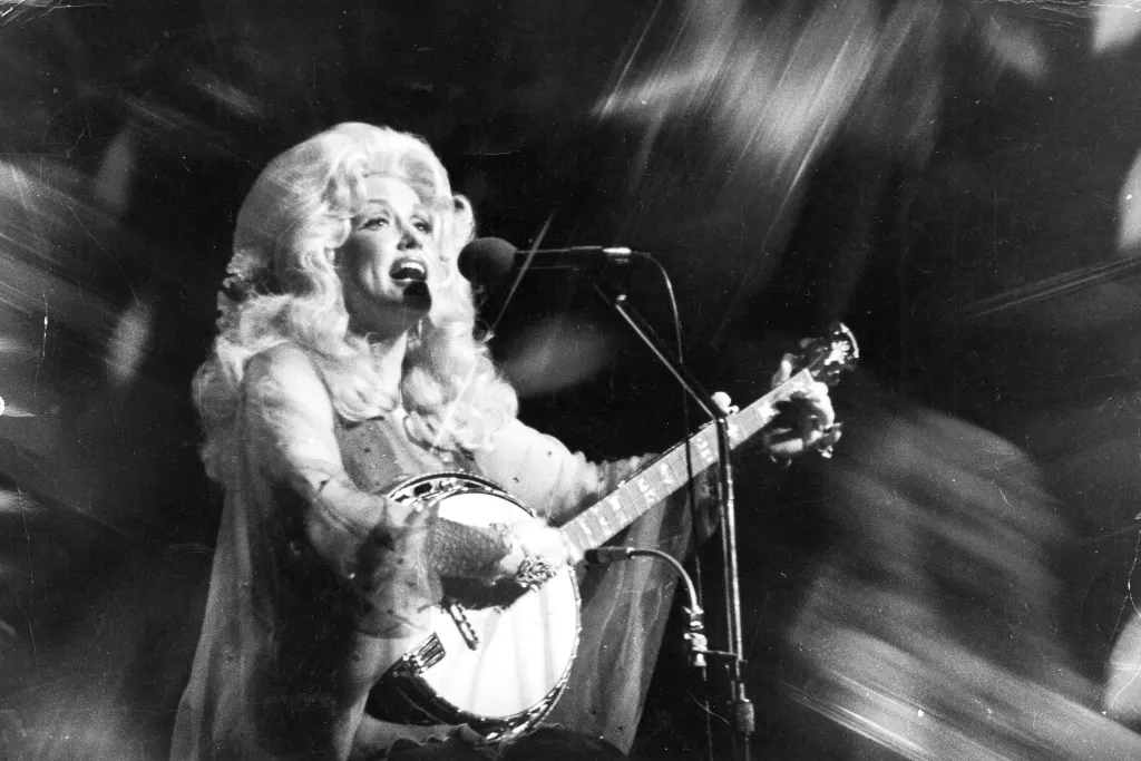 Dolly Parton sings into a microphone while playing a banjo.
