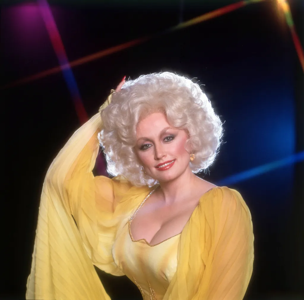 Dolly Parton smiling, wearing a yellow dress and a blonde wig, with a colorful striped background.