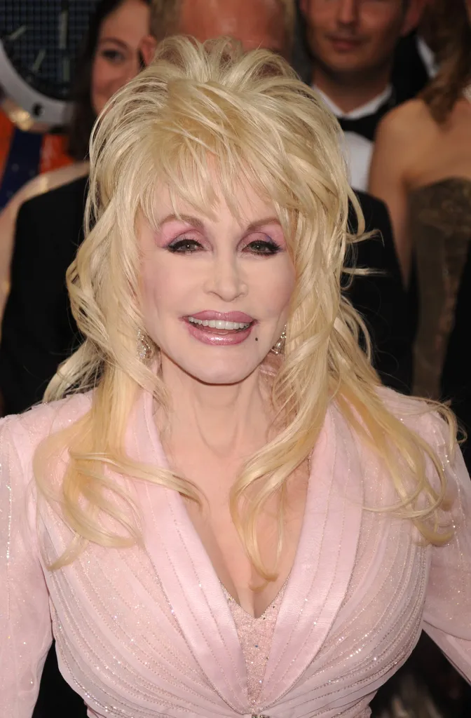 Dolly Parton smiling, wearing a light pink sparkling jacket and light pink lipstick.
