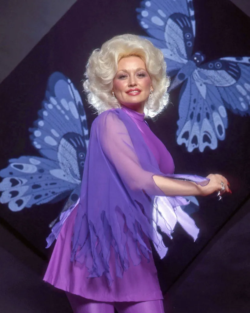 Dolly Parton smiling in a purple top and pants, with blue butterfly projections behind her.
