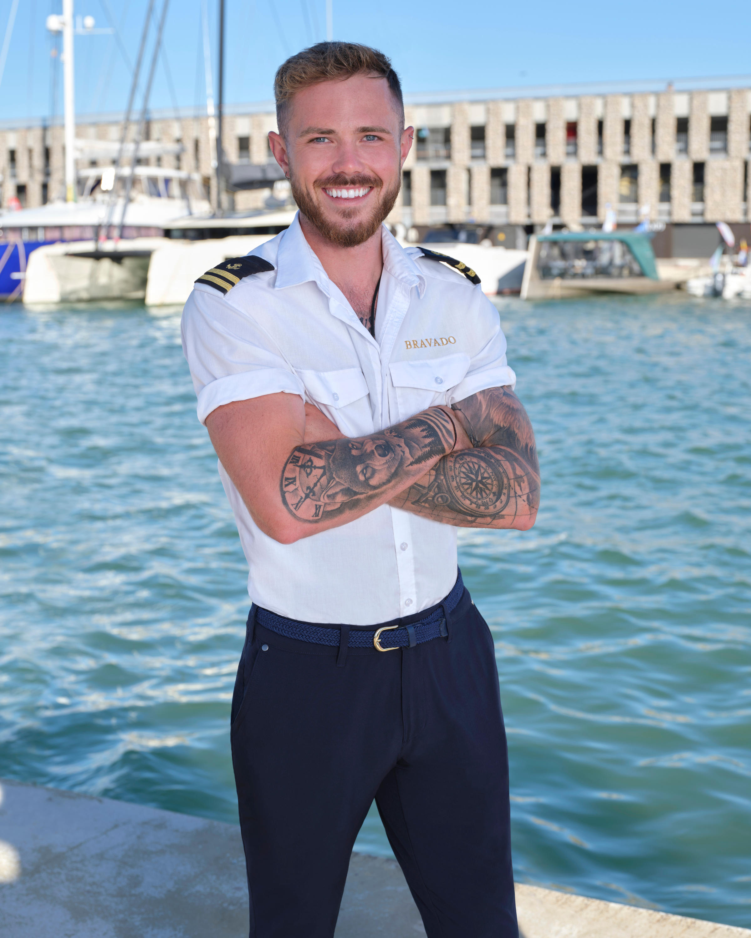 Nathan Gallagher, smiling in a uniform and crossing his tattooed arms, stands by the ocean.