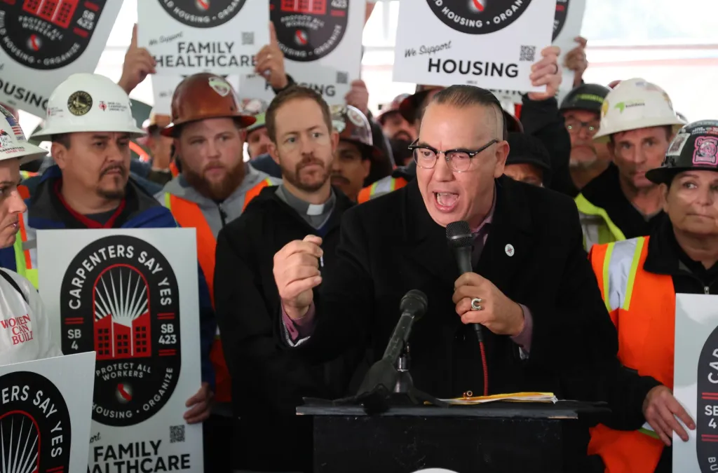 A man in glasses speaks into a microphone at a podium with a crowd of people holding signs behind him.