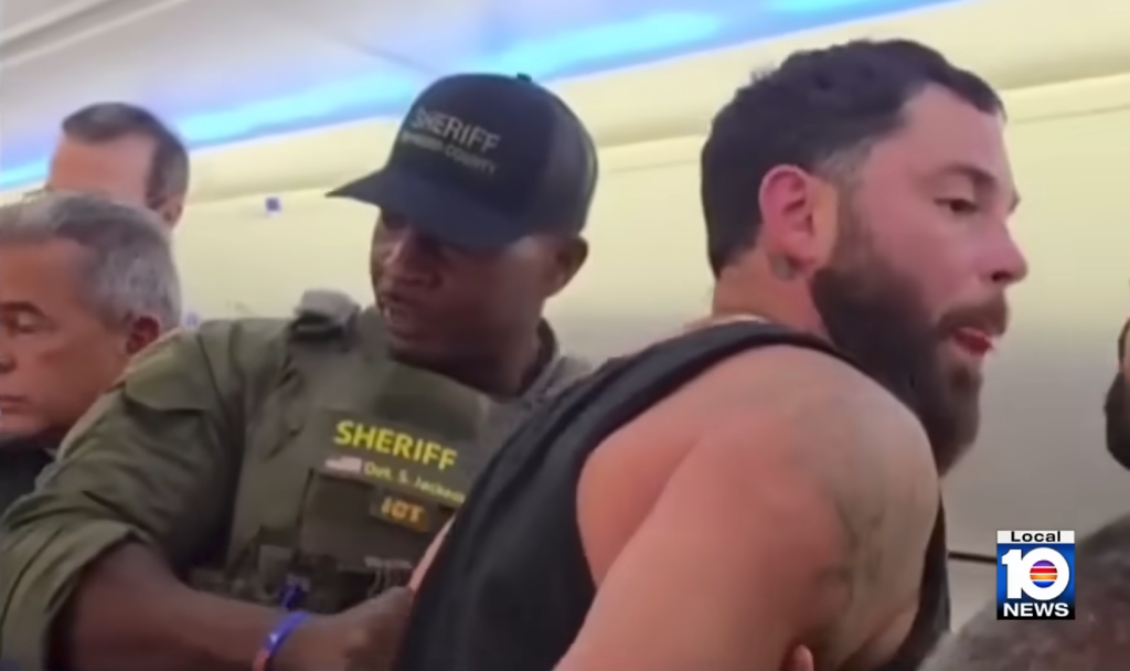 A police officer restraining a man in a black tank top inside an airplane.