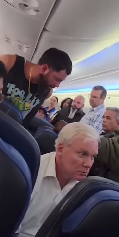 A man in a black tank top stands in the aisle of an airplane, looking down, while other passengers watch.