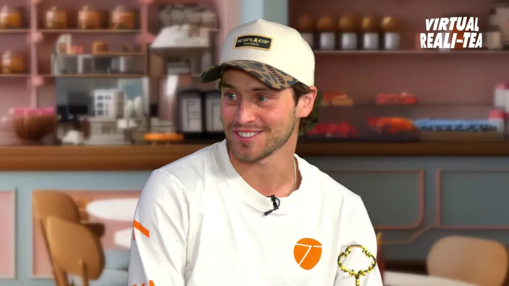 Liam Obergfoll, wearing a white shirt and a baseball cap, smiles in a cafe setting with the