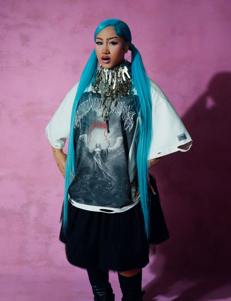 North West with bright blue hair, wearing an oversized graphic t-shirt, a key necklace, and a black skirt.
