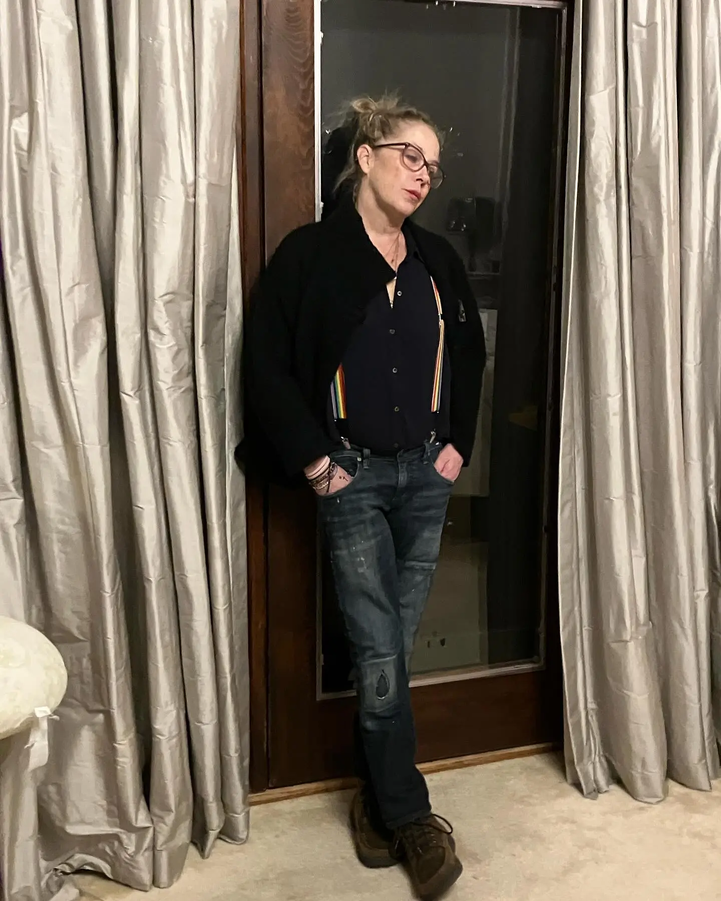Christina Applegate in a black shirt, jeans, and rainbow suspenders, leaning against a door.