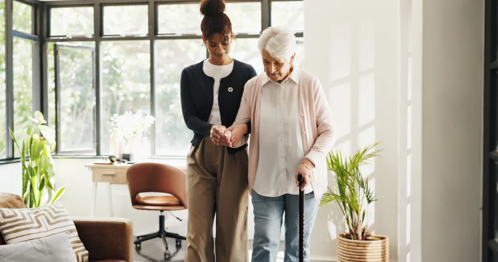 A nurse helping an elderly woman walk with a cane in a sunny room.