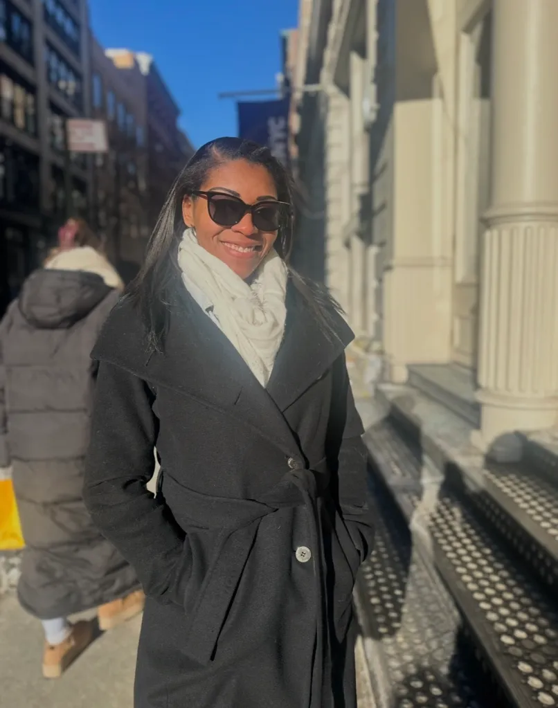 Jae Marie, smiling, wearing sunglasses and a scarf with a black coat, standing on a sunny street.