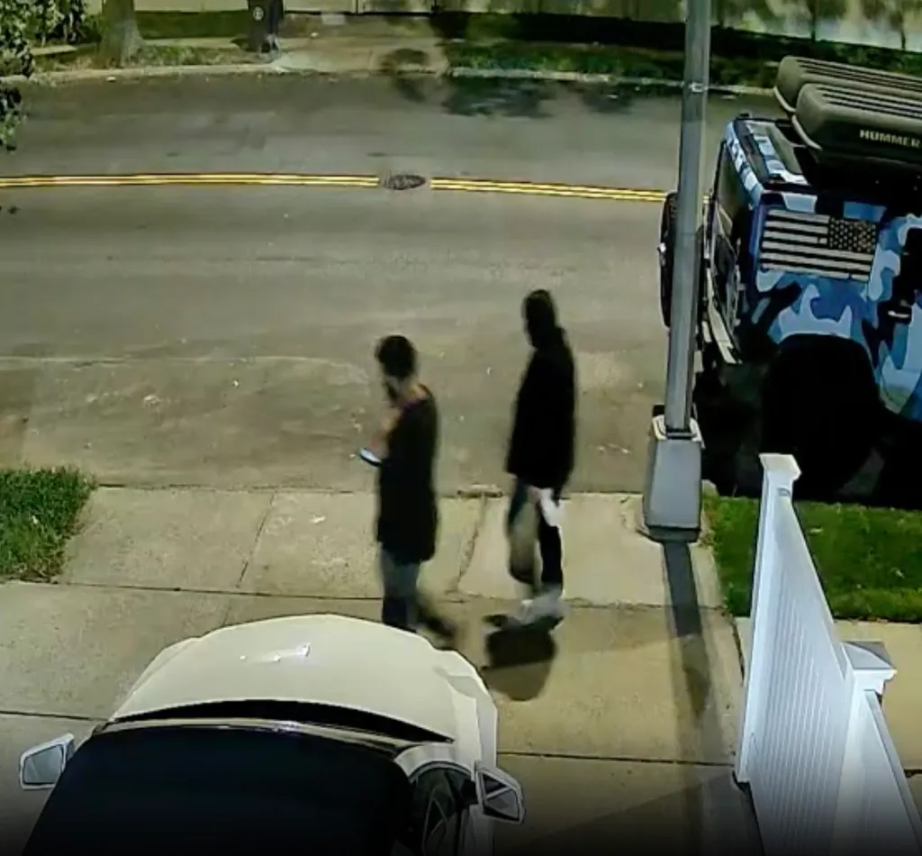 Security footage of two suspects walking on a sidewalk next to a vehicle.
