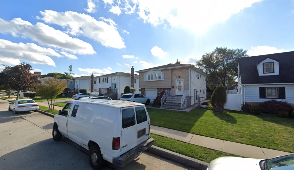 A suburban street with a white van in the foreground and a light brown two-story house with white trim and a white door with