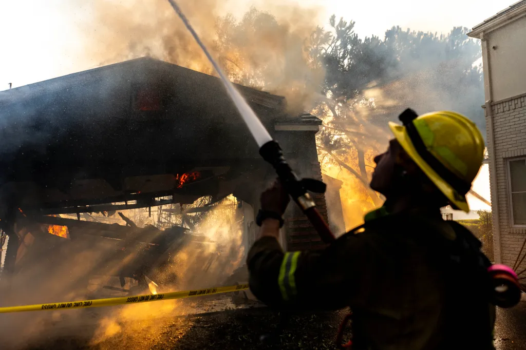 Firefighter battling the Palisades Fire, spraying water onto a burning structure with smoke and flames visible.