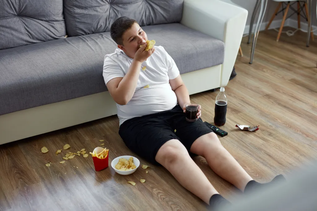 Overweight boy eating chips and drinking soda while sitting on the floor.