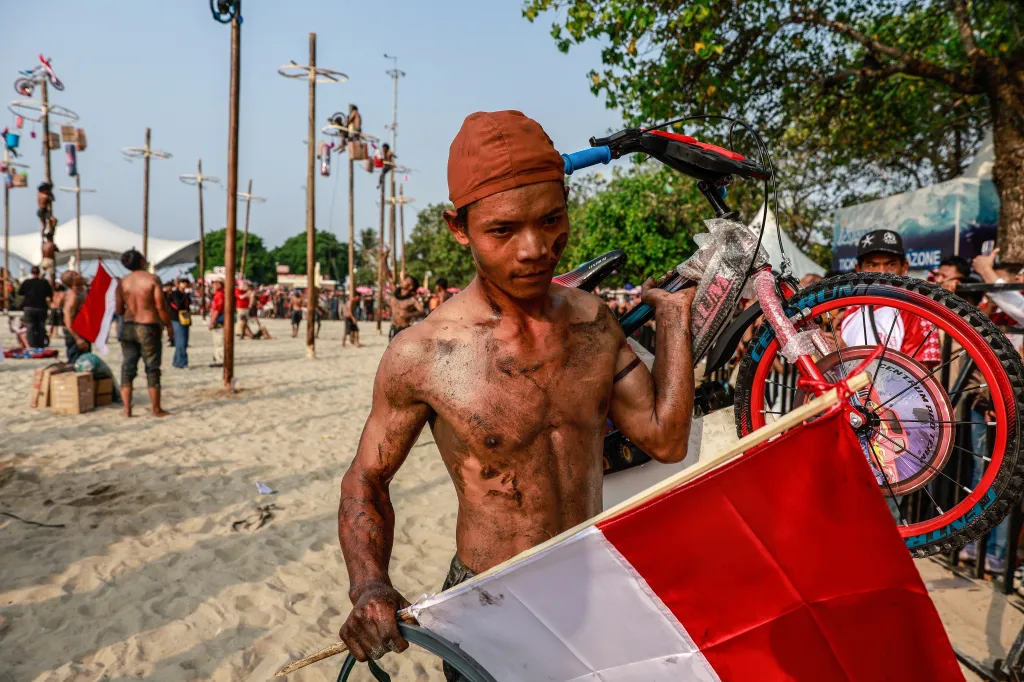 A shirtless participant, covered in grease, carrying a bicycle prize and the Indonesian flag after winning a greased pole race.
