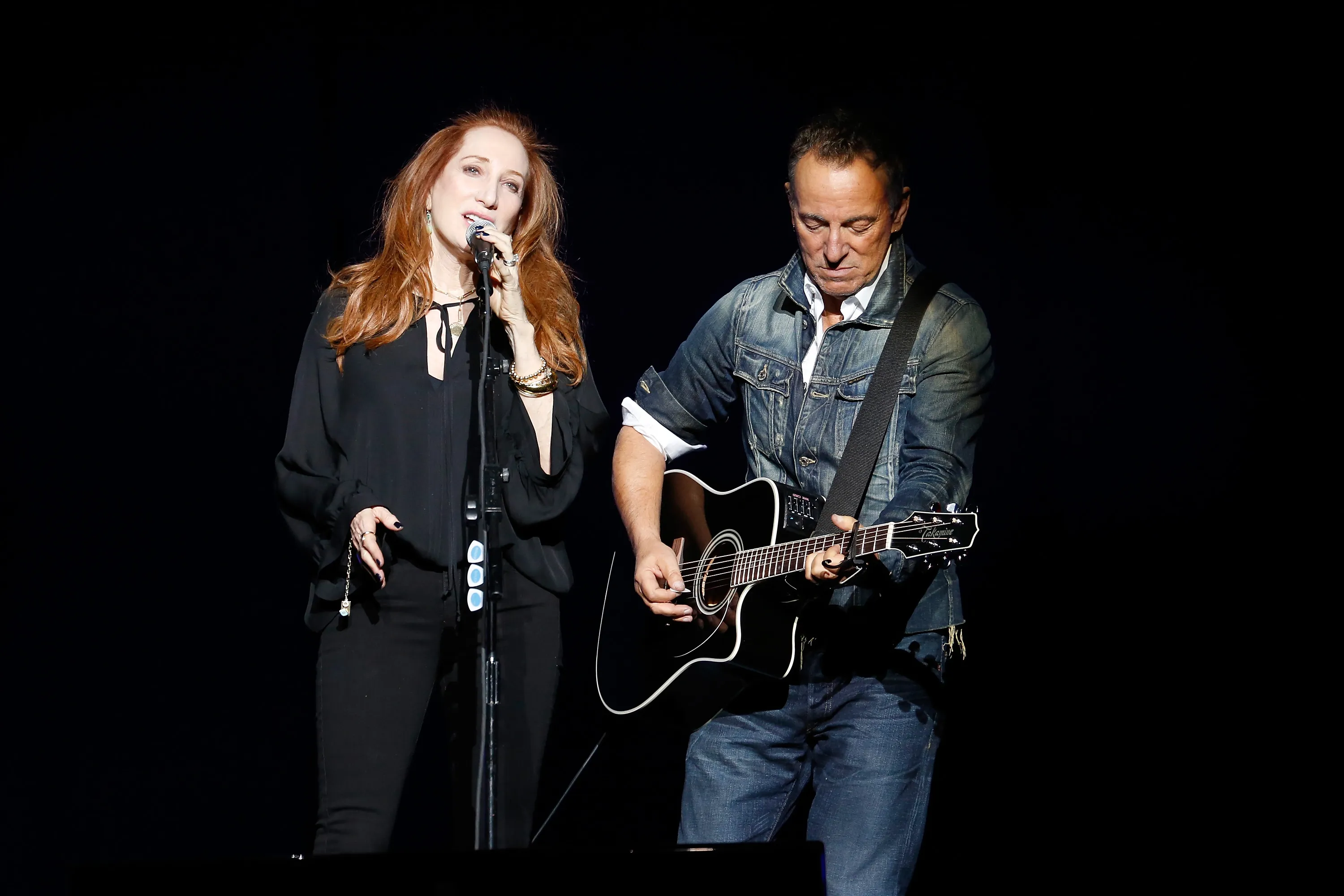 Patti Scialfa sings into a microphone while Bruce Springsteen plays an acoustic guitar on stage.