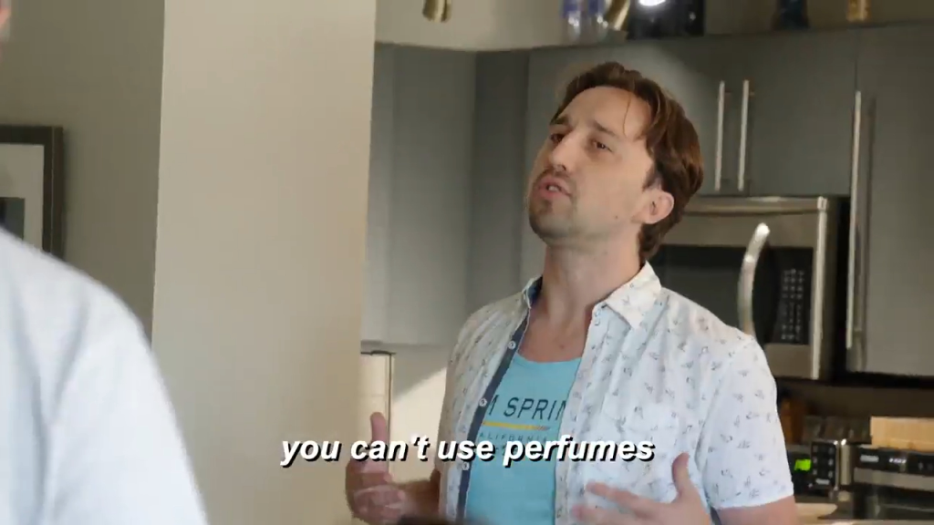 A man in a light blue shirt with a collared, patterned short-sleeved shirt over it, talking to someone off-screen with the caption