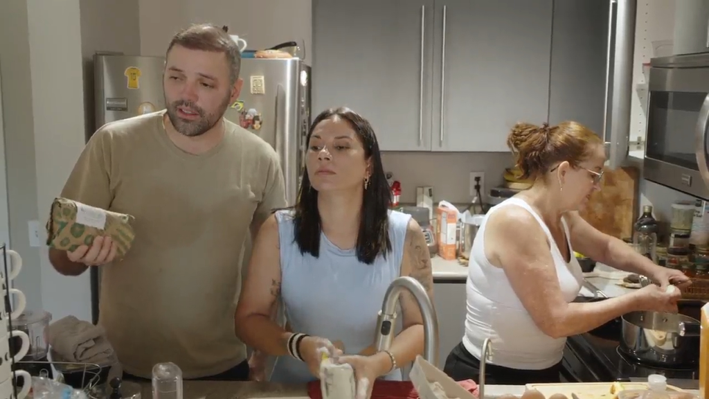 Three people in a kitchen, a man in a tan shirt holding a package, a woman in a light blue top washing something, and another woman in a white tank top cooking at the stove.