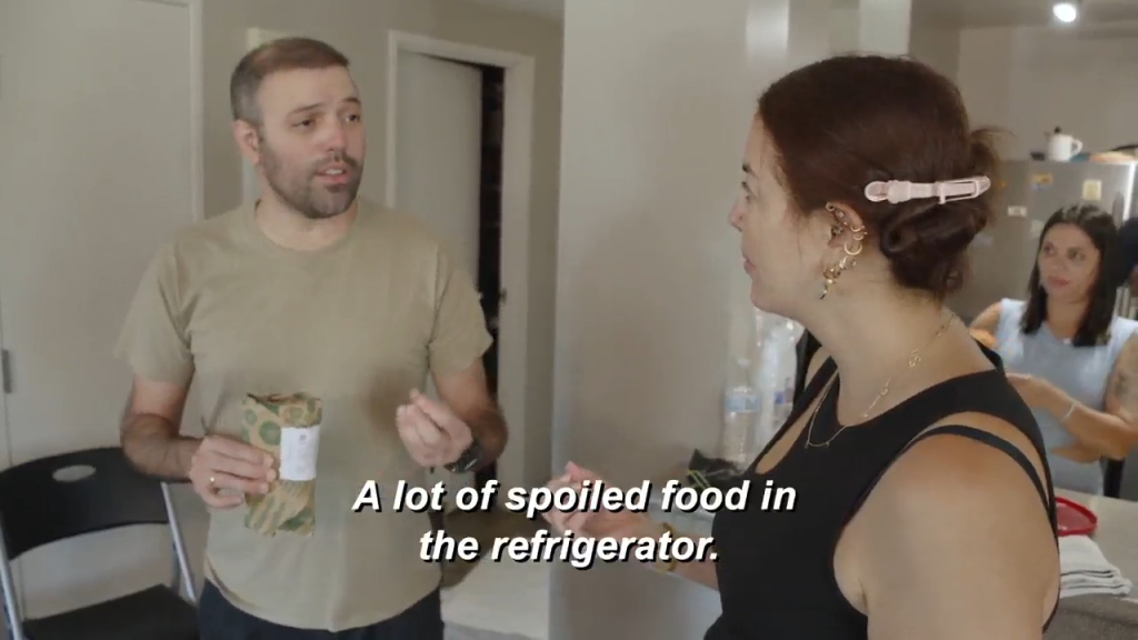 A man holding a wrapped package talks to a woman with a hair clip, while another woman is visible in the background.