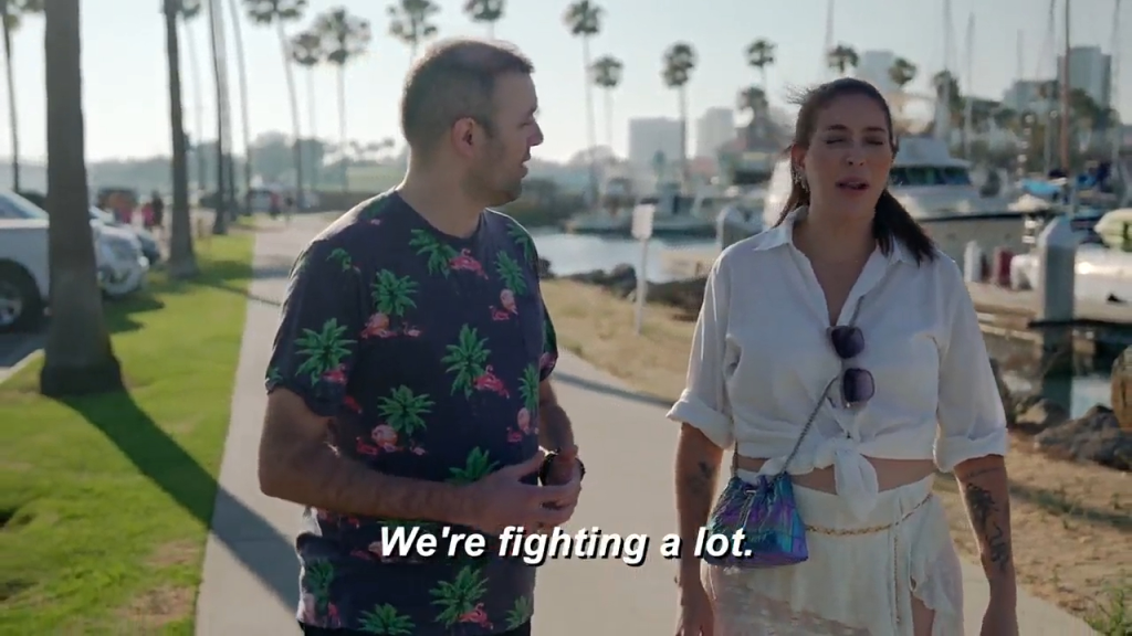 A man and a woman walk along a boardwalk beside a marina, the woman says,