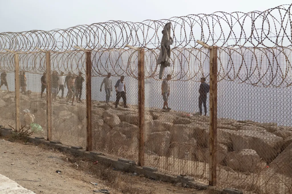 People walk on rocks behind a barbed wire fence with the sea in the background.