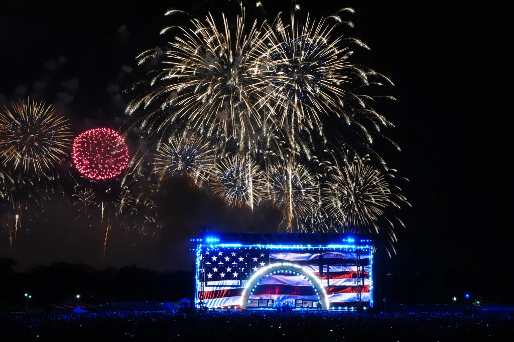 Fireworks light the sky above a stage displaying an American flag for an Independence Day celebration.
