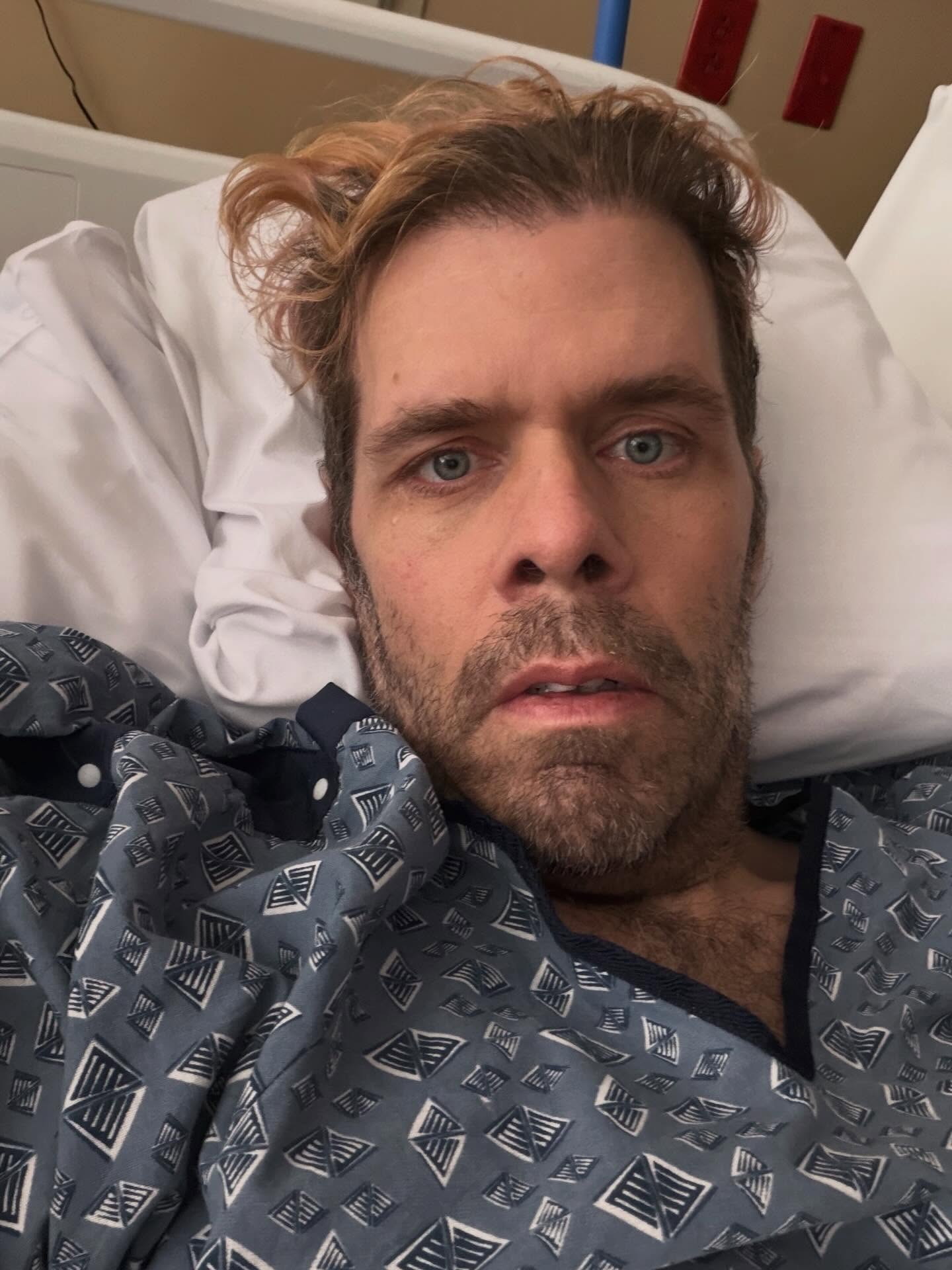 Perez Hilton lying in a hospital bed wearing a patient gown.