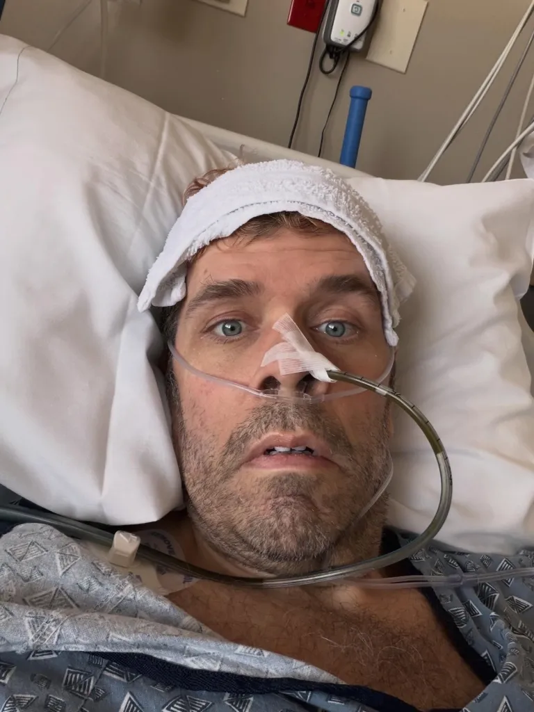 Perez Hilton in a hospital bed with a white towel on his head, and a nasal cannula and feeding tube in his nose.