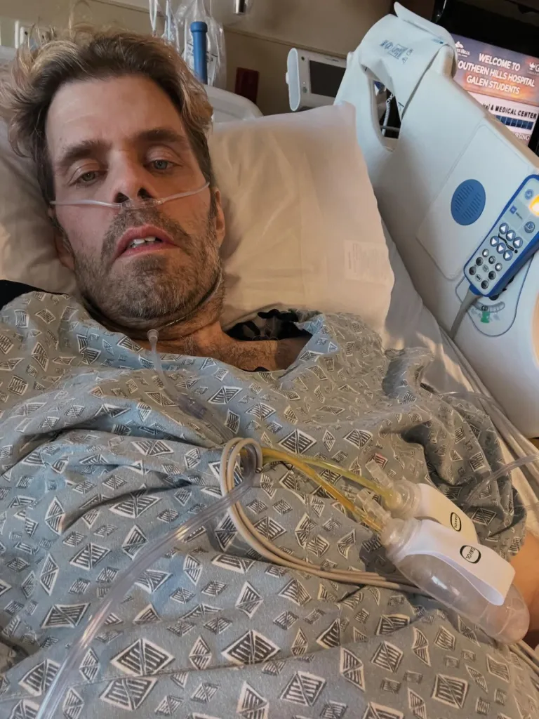 Perez Hilton in a hospital bed with tubes in his nose and what appears to be a wound drainage system.