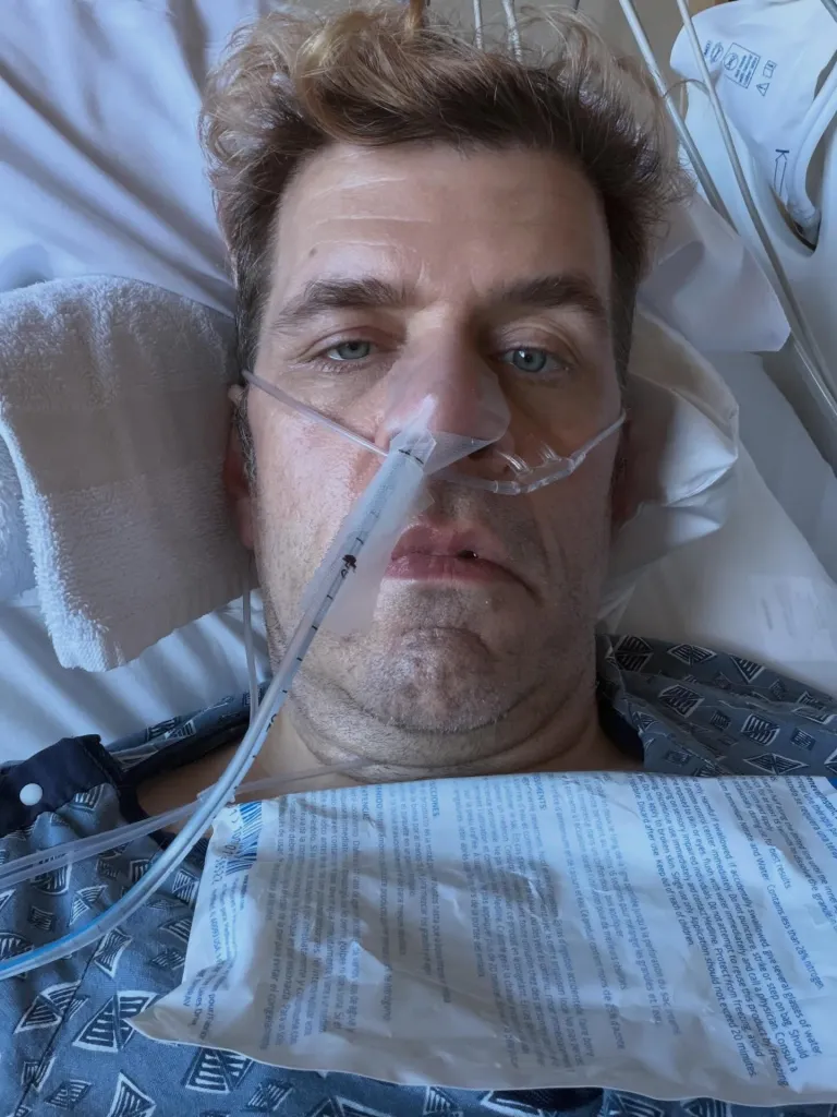 Perez Hilton lying in a hospital bed with a nasal cannula and a cold compress on his chest.