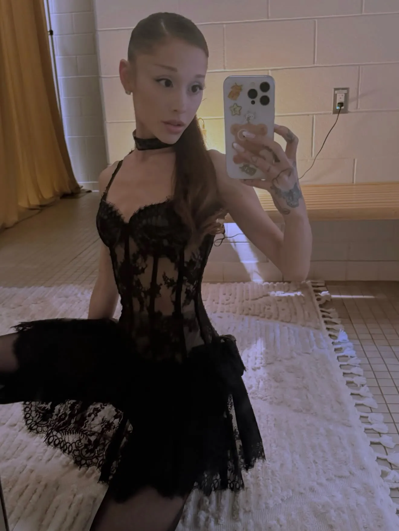 Ariana Grande in a black lace corset and skirt taking a selfie.