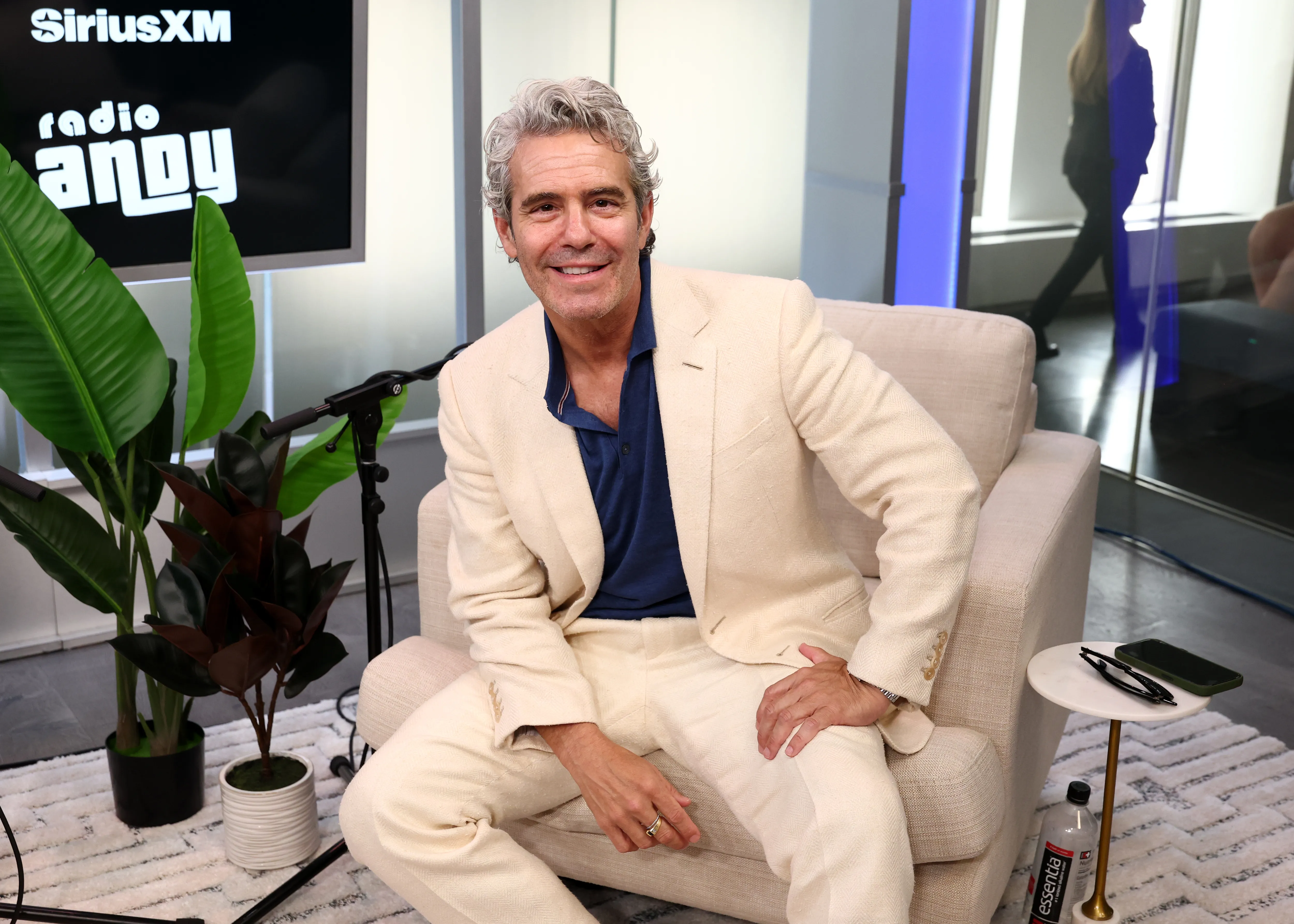 Andy Cohen smiles while sitting on a light-colored armchair with a