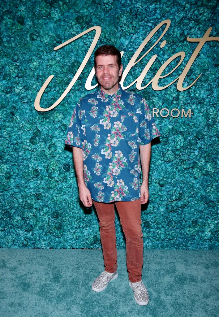 Perez Hilton in a floral shirt and brown pants posing against a teal floral wall at the grand opening of Juliet Cocktail Room.